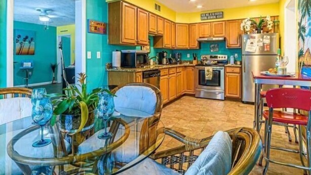 A kitchen with wooden cabinets, a dining area with wicker chairs, and a ceiling fan. - Port Canaveral/Space X- steps from beach 2-bedroom colorful unit w/ AC-sleeps 8 (Cocoa Beach)