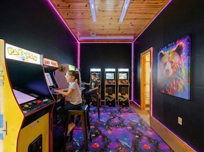 A room with arcade machines, a bear painting, and a space-themed carpet. - Heated Pool, Sleeps 30, 6,000 sq ft. Family Cabin, Arcade, Pool Table, Bunk Room (Broken Bow)