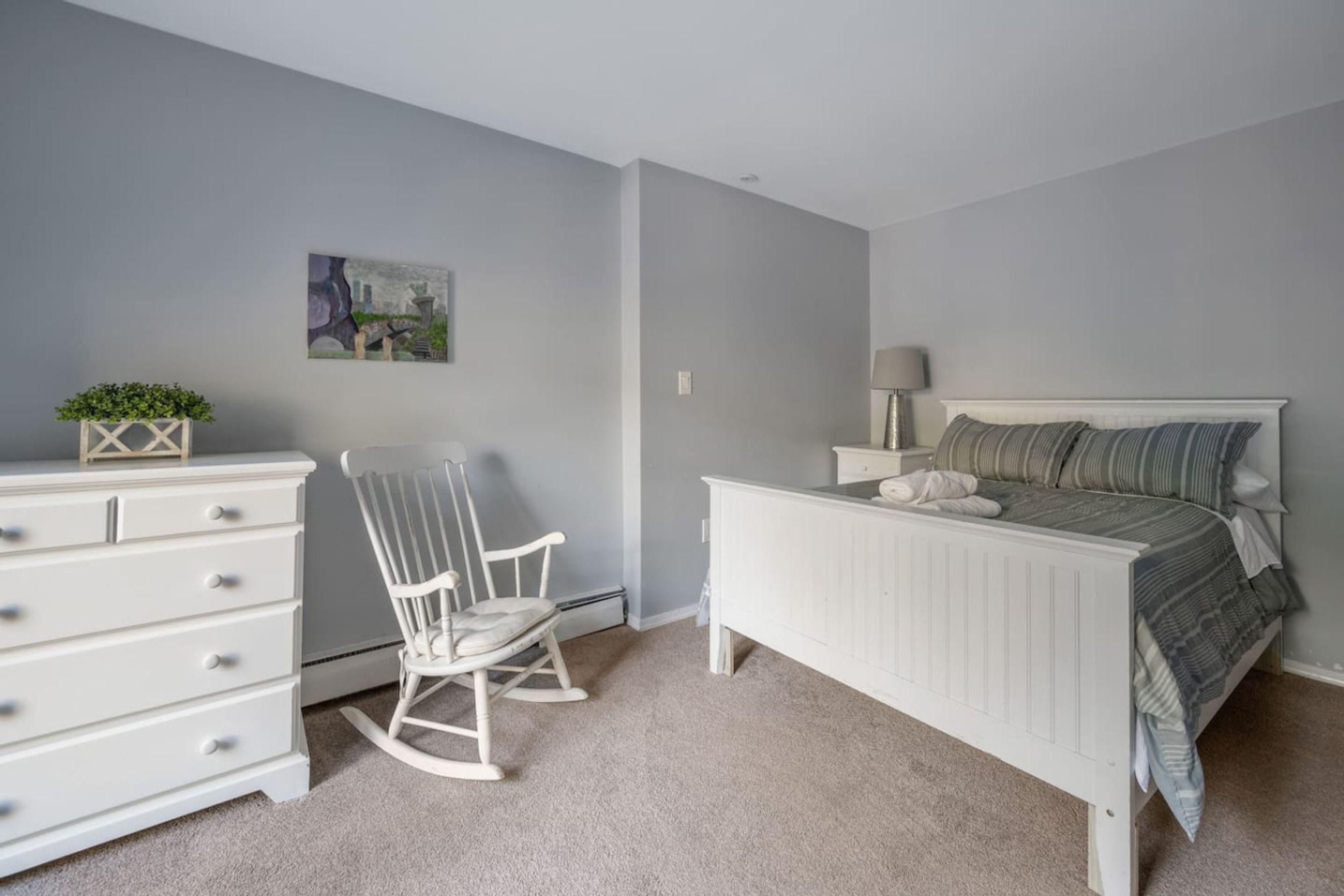 Un dormitorio con cama, cómoda, mecedora y un cuadro en la pared.