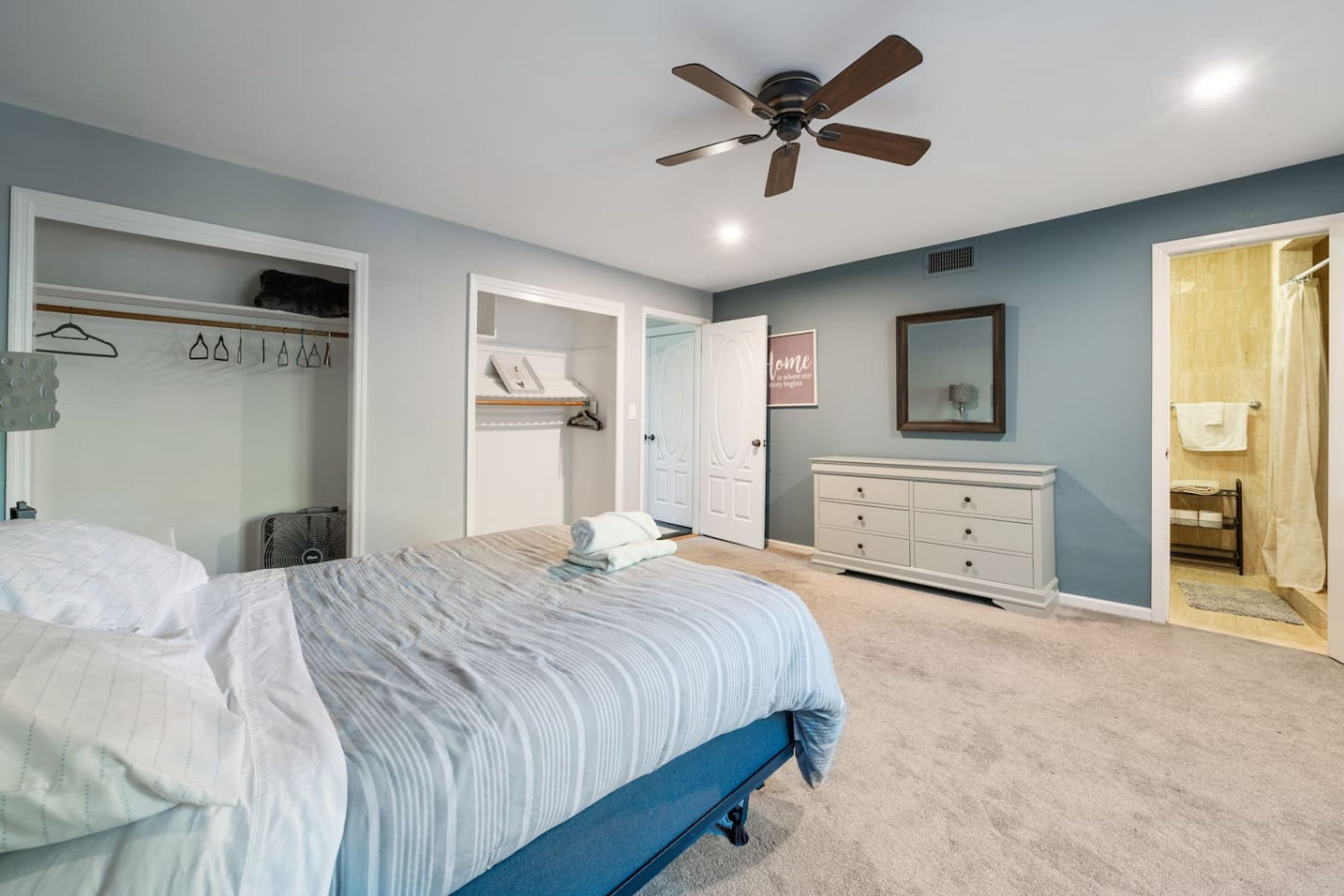 A bedroom with a bed, a ceiling fan, a walk-in closet, a dresser, and a shower area.