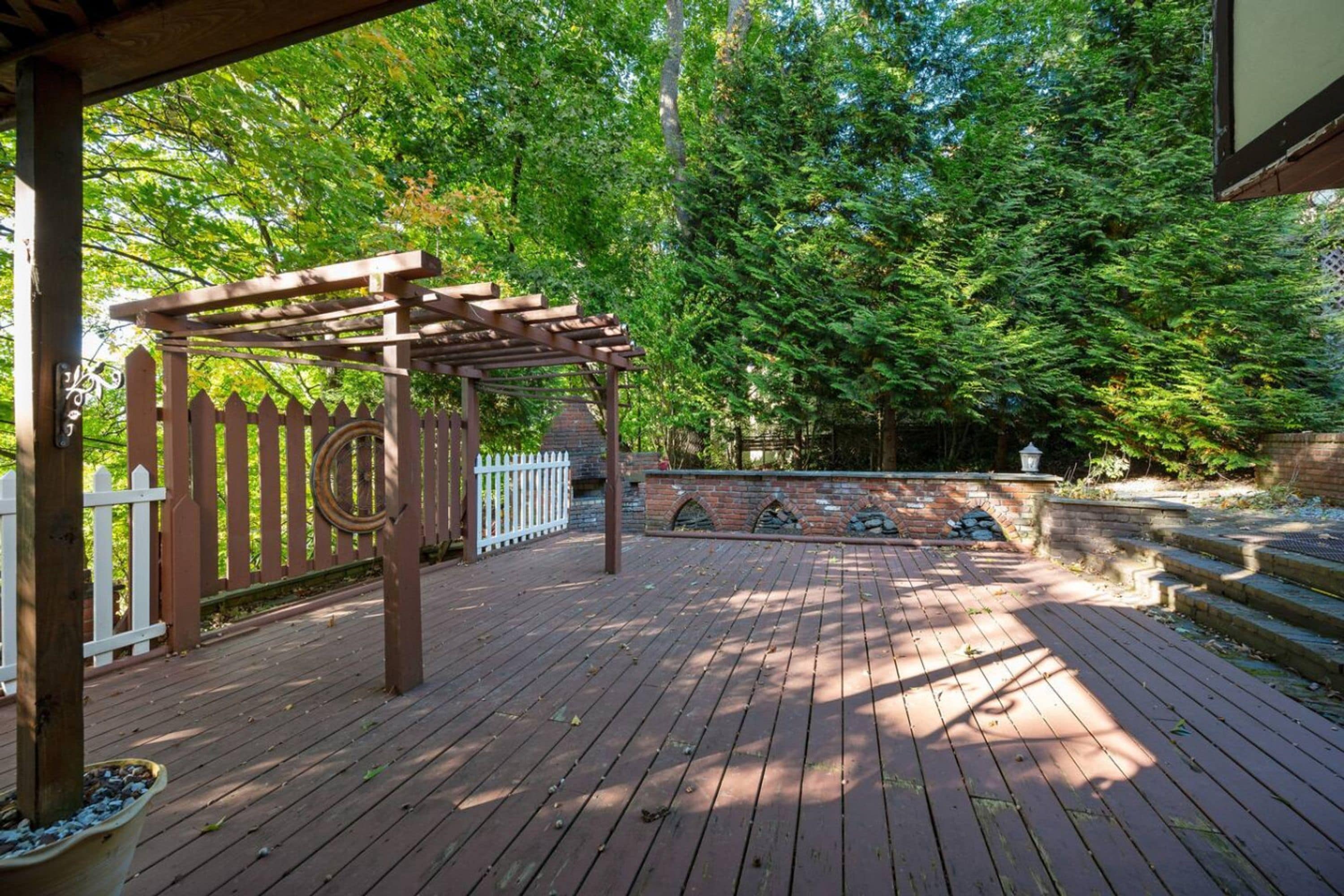 A wooden deck with a pergola, surrounded by a white picket fence and a brick wall.
