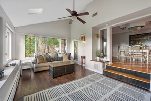 Ein geräumiges Wohnzimmer mit großem Fenster, Deckenventilator, Essbereich mit Tisch und Stühlen sowie einer Treppe zu einer oberen Ebene.