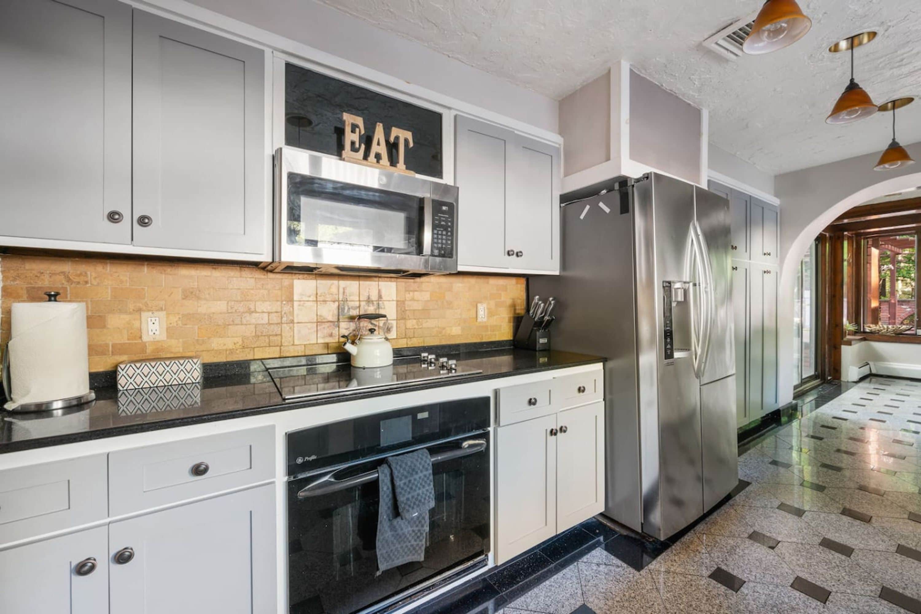 Een moderne keuken met roestvrijstalen apparatuur, een magnetron en een koelkast. Er is een aanrecht met een waterkoker en een keukenrol. De keuken heeft witte kastjes en een betegelde spatwand.