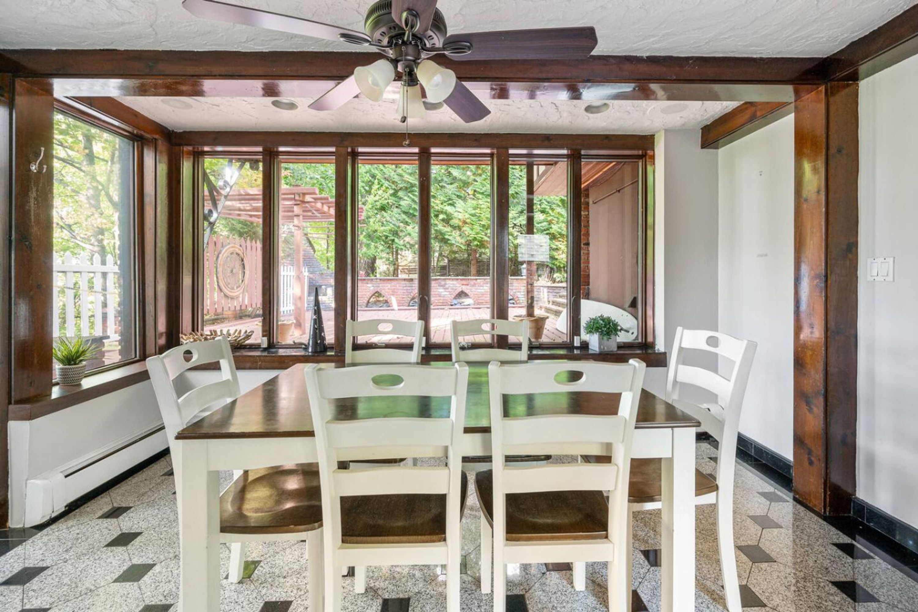 Uma sala de jantar com uma mesa grande e cadeiras, um ventilador de teto e janelas amplas com vista para o exterior.