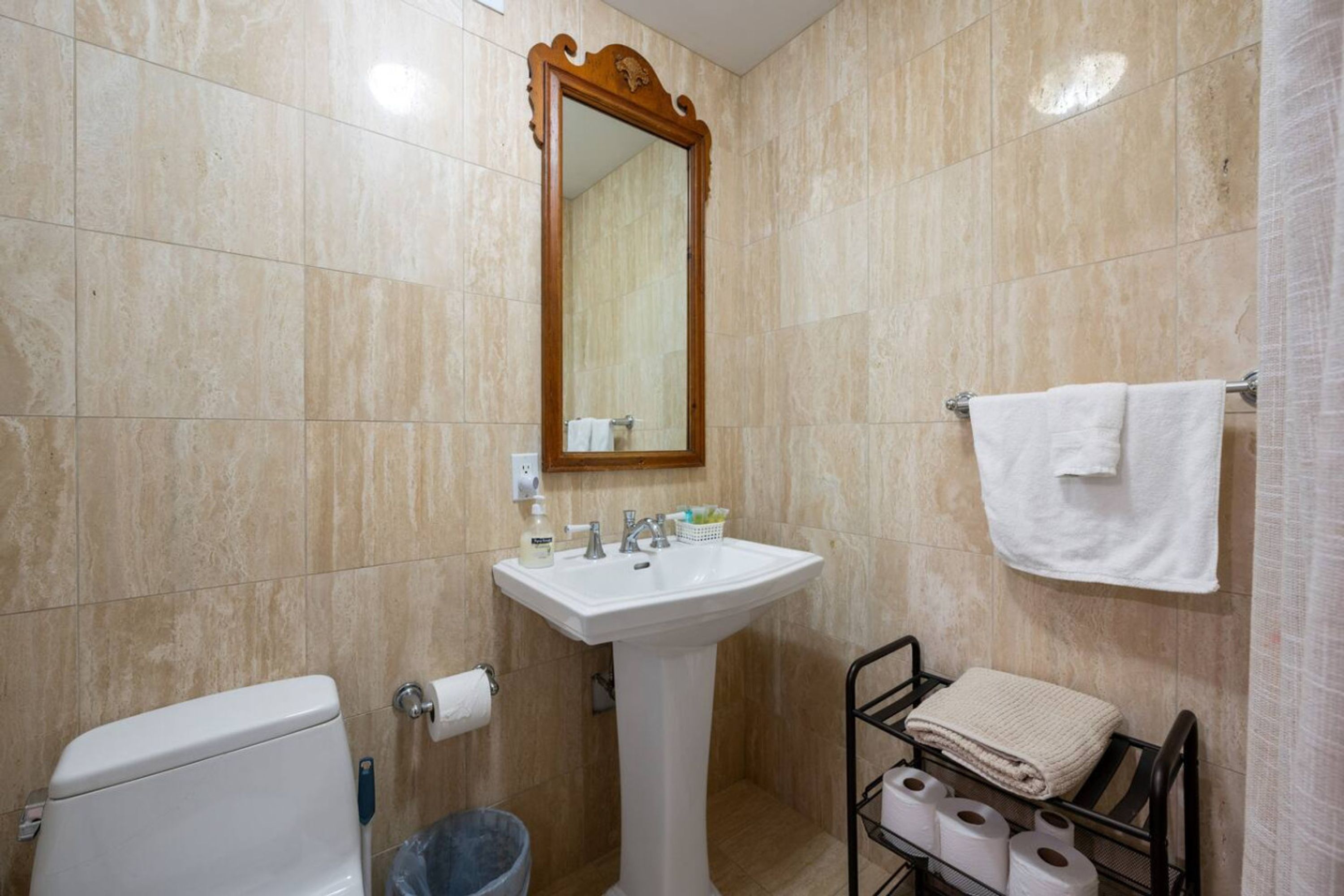 Un baño con un lavamanos blanco de pedestal, un espejo con marco de madera, un inodoro, un cubo de basura y un toallero con toallas.