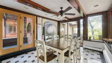 A dining area with a wooden table and chairs, a ceiling fan, and large windows offering views of the outdoors.