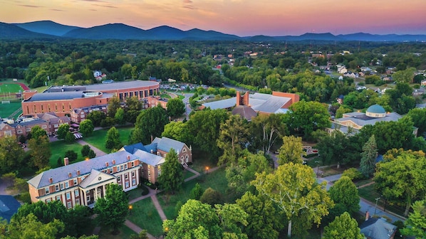An aerial view of a campus with multiple buildings, including a large brick structure and several smaller houses, surrounded by greenery and a mountainous backdrop.