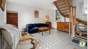 Un salon avec un canapé bleu, une table basse en bois et un sol à carreaux. On aperçoit un escalier en bois à l’arrière-plan.