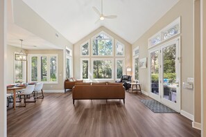 A spacious living room with a large brown sofa, a dining area with a table and chairs, and large windows providing natural light.