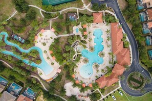 Vista aérea de um resort com várias piscinas, um lago serpenteante e um edifício central com telhado de telhas vermelhas.
