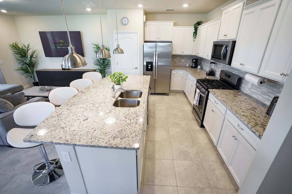 Una cocina moderna con armarios blancos, encimeras de granito, electrodomésticos de acero inoxidable y taburetes de bar.