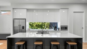 A modern kitchen with a large island, bar stools, a refrigerator, a microwave, and a view of greenery through the window. - Belle Escapes - Henley Beach House (Henley Beach)