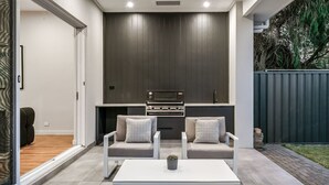 A modern kitchen with dark cabinetry, a built-in oven, and a sink. There are two grey armchairs with patterned cushions and a white coffee table with a plant. - Belle Escapes - Henley Beach House (Henley Beach)
