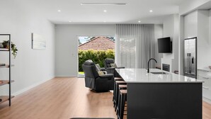 A modern kitchen with a central island, a dining area with a sofa, and a view of a house through a large window. - Belle Escapes - Henley Beach House (Henley Beach)