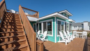 Een houten terras met witte Adirondack-stoelen, een overdekte patio en een trap naar een groen huis met witte sierlijsten.