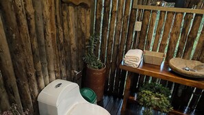 A rustic bathroom with a wooden floor, a white toilet, a wooden shelf with a basin, and a potted plant.