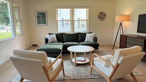 A living room with a sectional sofa, two armchairs, a coffee table, and a floor lamp. There are two windows with blinds, a painting on the wall, and a rug on the floor. - Spacious 3BR 2BA Gem Close to Downtown, 30 day min (Eau Claire)