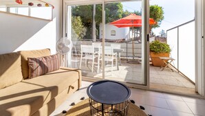 A modern living room with a sofa, a small round table, and a sliding glass door leading to an outdoor patio area with a white table and chairs, a red umbrella, and a potted plant.
