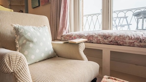 A cozy room with a striped armchair, a cushioned bench with a floral cushion, a small table with a book and glasses, and a view of a balcony with a railing. - 11b Market Cross Place, Aldeburgh (Aldeburgh)