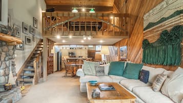 A spacious living area with a high ceiling, wooden beams, a staircase, and a kitchen in the background.