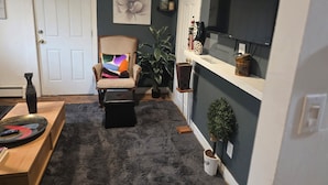 Un salon avec un mur foncé, une porte blanche, une chaise en bois avec un coussin coloré, une petite table avec un bol et une télécommande, et une plante en pot.
