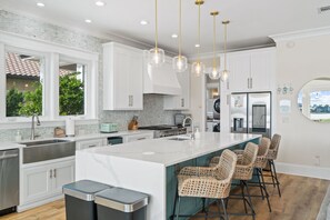 Una cucina moderna con mobili bianchi, un'isola grande con sgabelli da bar, elettrodomestici in acciaio inossidabile e illuminazione a sospensione.