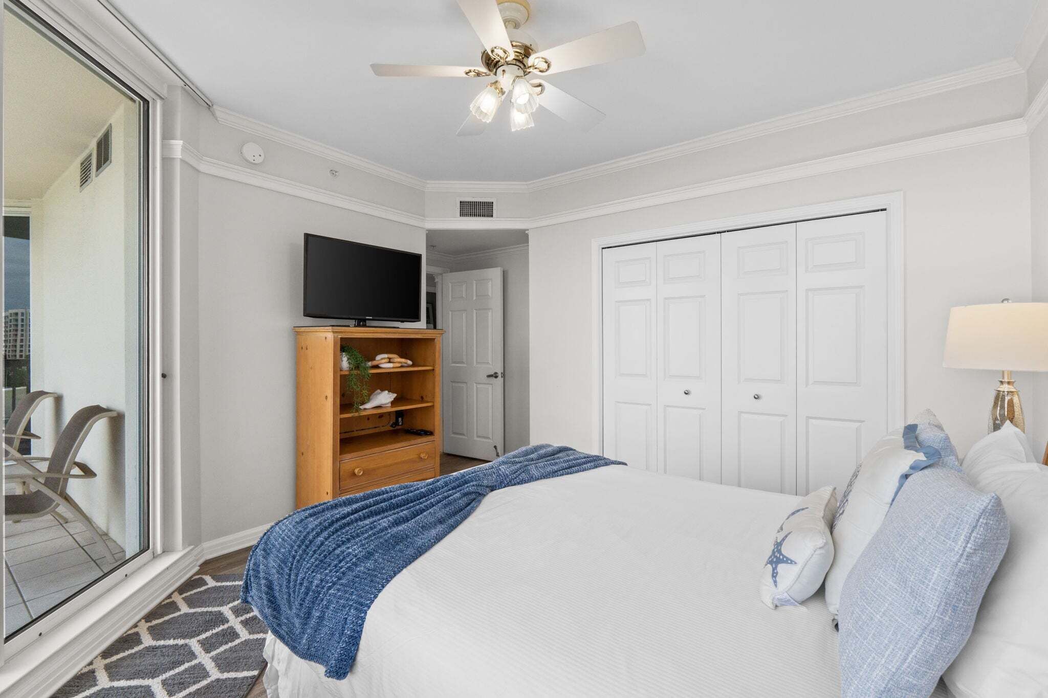 A bedroom with a bed, a television on a wooden stand, a ceiling fan, and a door.