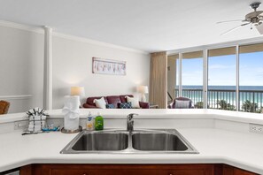 Una cocina con un lavamanos doble, una ventana con vista al océano, un ventilador de techo y un comedor con sofá y mesa.