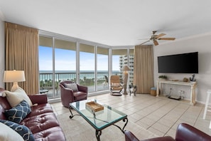 A spacious living room with a large window, a flat-screen TV, a ceiling fan, and a comfortable seating area.