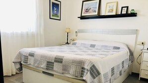 A neatly made bed with a quilt, a bedside lamp, and a shelf with framed pictures. - ROCMAR Apartment opposite the port of Roses. (Roses)