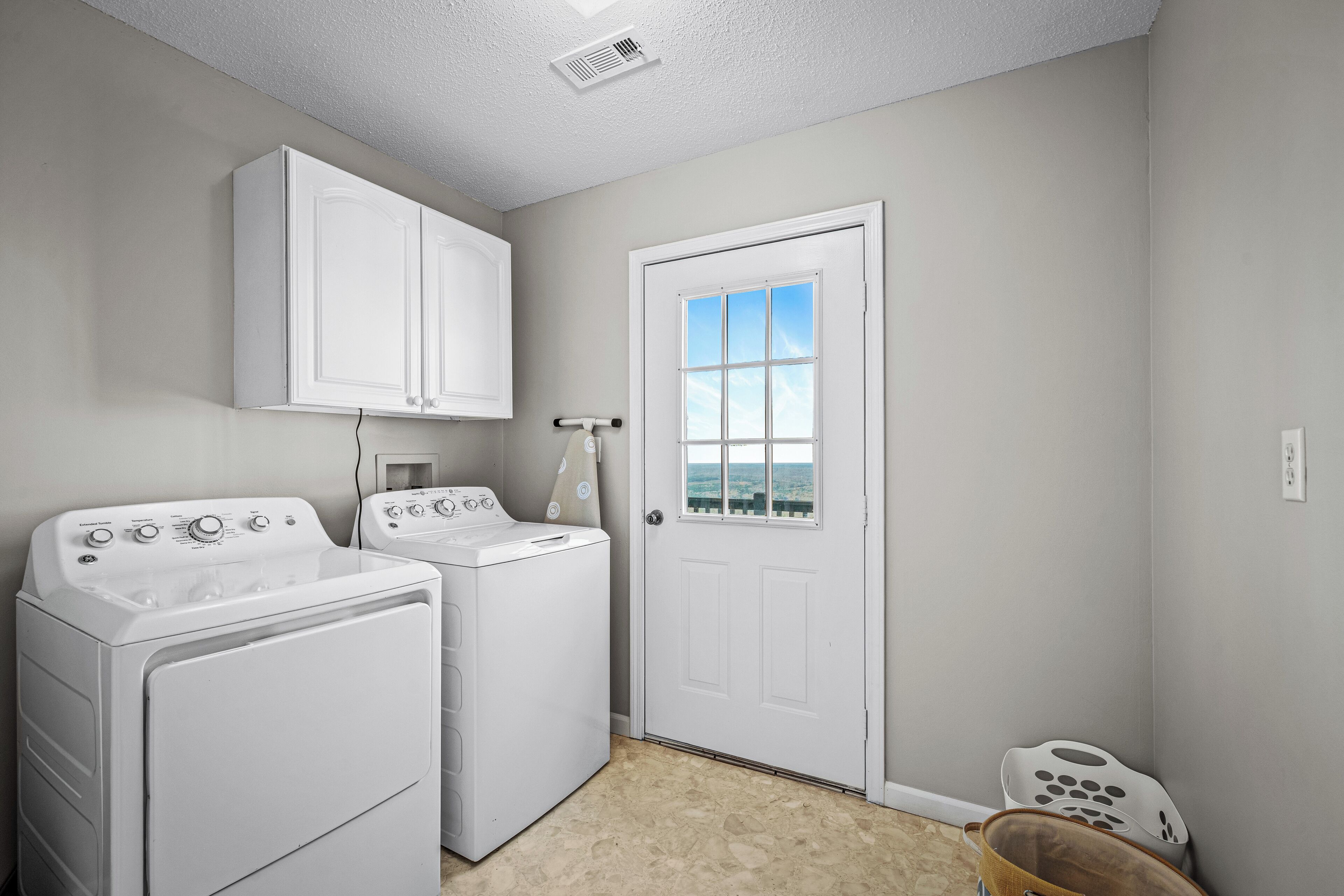 A laundry room with two white washers and dryers, a white cabinet above the dryers, a white door with a window, and a small laundry basket.