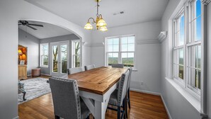 A dining area with a wooden table and grey upholstered chairs, large windows, and a ceiling fan. - Best View Above Mentone - High Brow House - Elevate to Escape (Mentone)