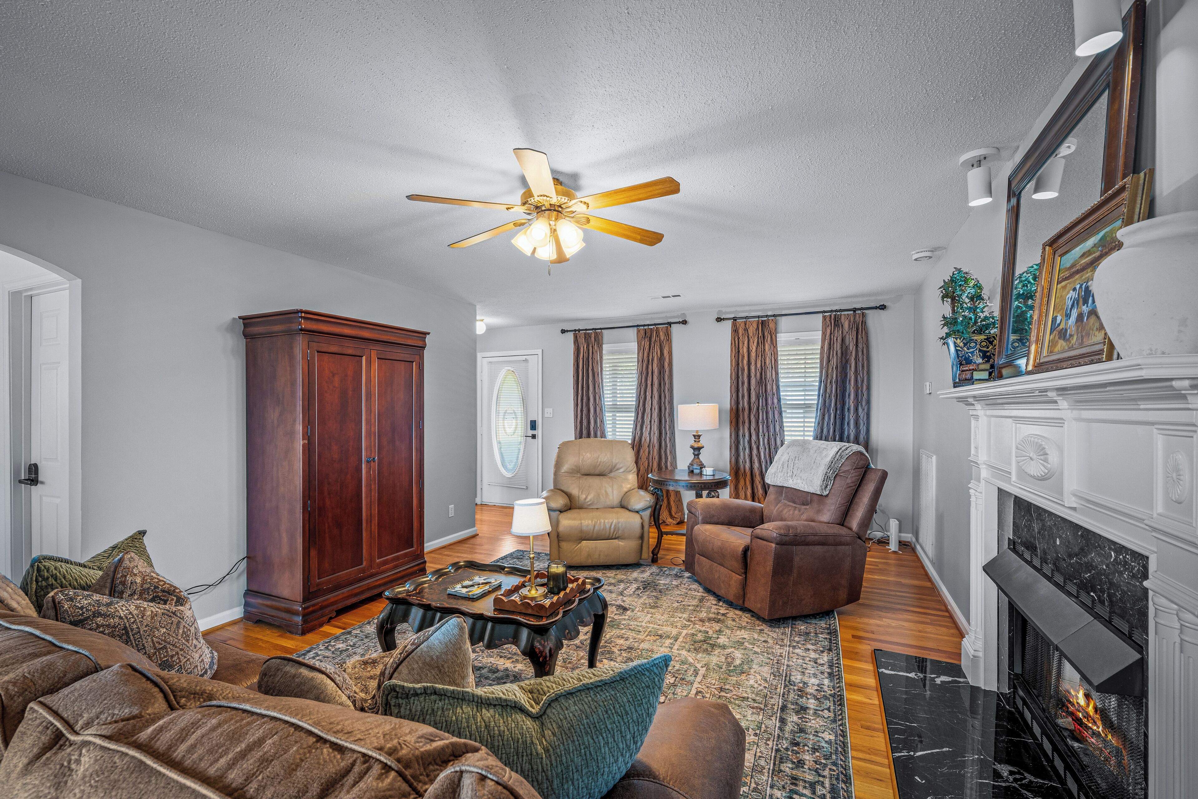 A spacious living room with a fireplace, a ceiling fan, and a large wooden cabinet.