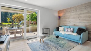 A modern living room with a glass coffee table, a blue sofa, and a view of a park through large sliding glass doors. - Central Parc 2 by Interhome (Crans-Montana)