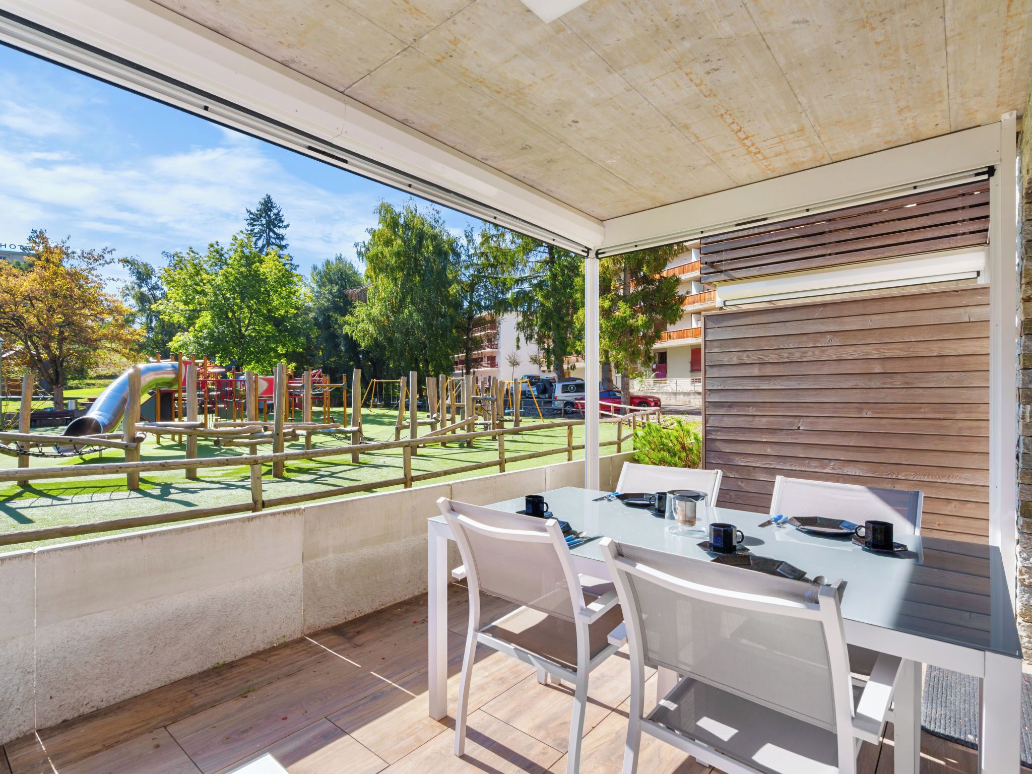 A covered outdoor area with a table and chairs, a playground in the background, and a view of a residential area.