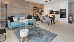 A modern living room with a blue sofa, a glass coffee table, and a dining area with chairs. - Central Parc 2 by Interhome (Crans-Montana)