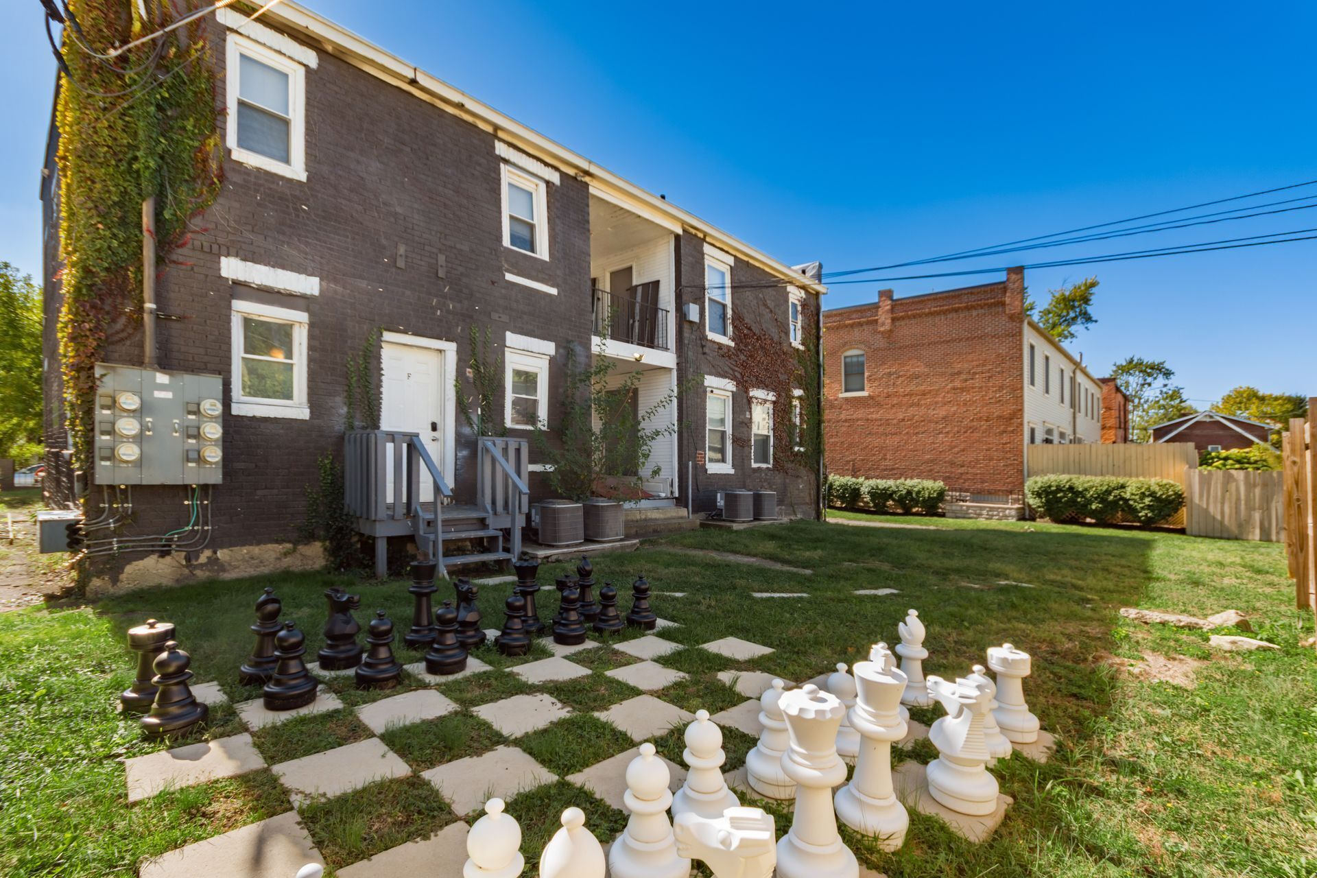A residential area with a large chessboard set up on the lawn, a house with a brick facade, and a utility box.