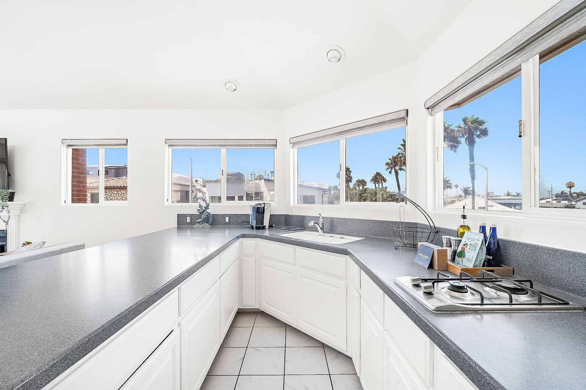 Una cocina con armarios blancos, encimera de granito, estufa a gas y ventanas amplias que ofrecen vistas a palmeras y edificios.