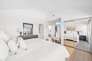 A hotel room with a large bed, a bedside table with a lamp, a mirror, and a television. - Luxury 4BR Retreat | AC | Balcony & Rooftop Deck with Ocean Views! (Newport Beach)