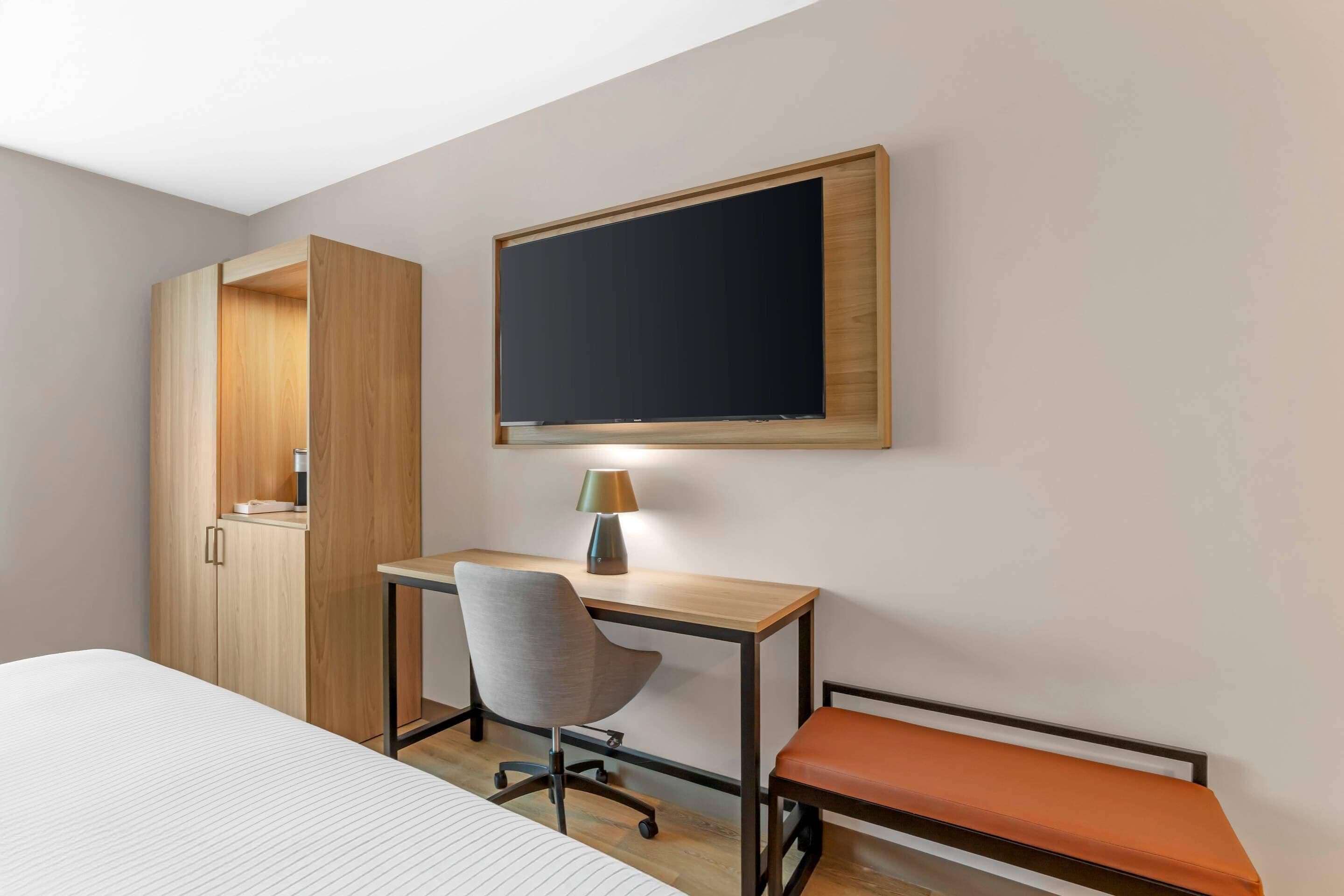A hotel room with a bed, a desk with a chair, a television mounted on the wall, and a wardrobe.