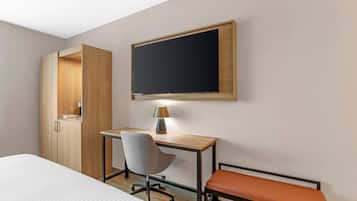 A hotel room with a bed, a desk with a chair, a television mounted on the wall, and a wardrobe.