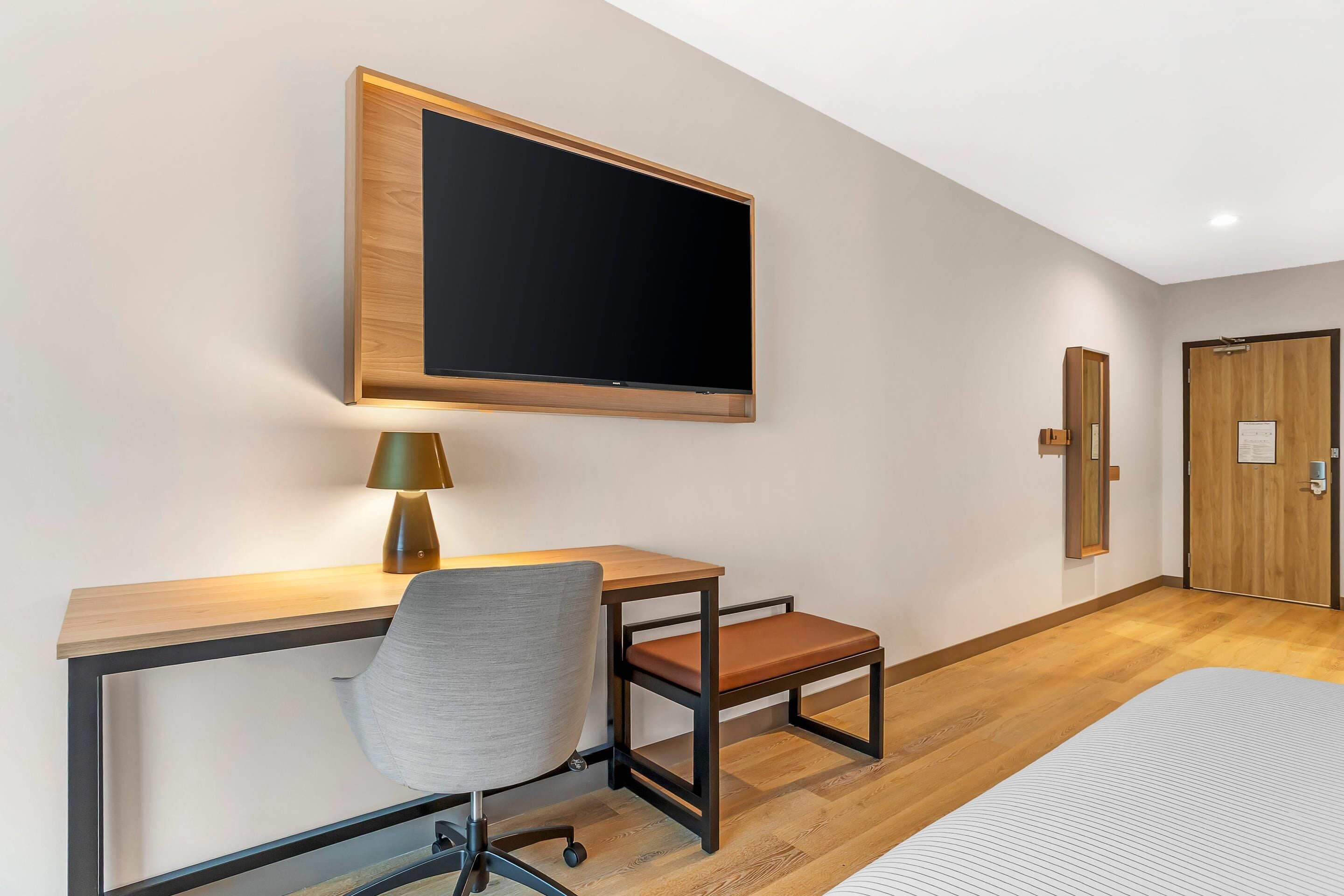 Ein Hotelzimmer mit einem Bett, einem Schreibtisch mit Stuhl, einem an der Wand befestigten Fernseher und einer Tür.