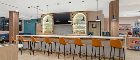 Eine moderne Bar mit einer langen Theke, mehreren Barhocker, Pendelleuchten und einer Auswahl an Snacks und Getränken.