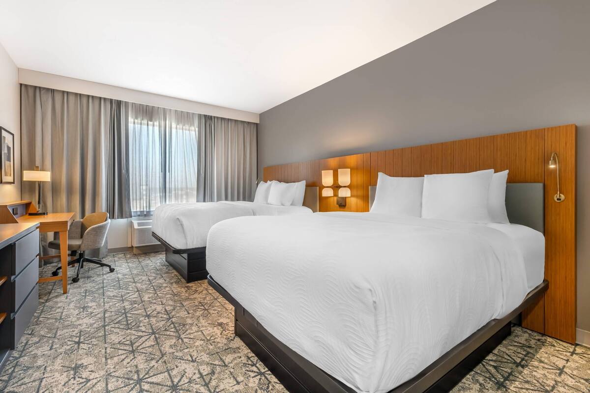 A hotel room with two beds, a desk, a chair, and a large window with curtains.