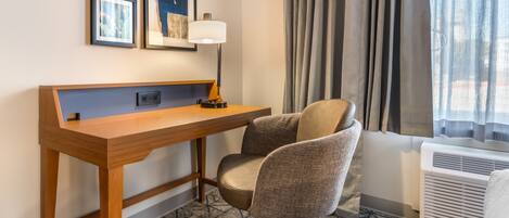 A hotel room with a wooden desk, a cushioned office chair, a wall-mounted lamp, framed pictures, a window with curtains, and an air conditioning unit.