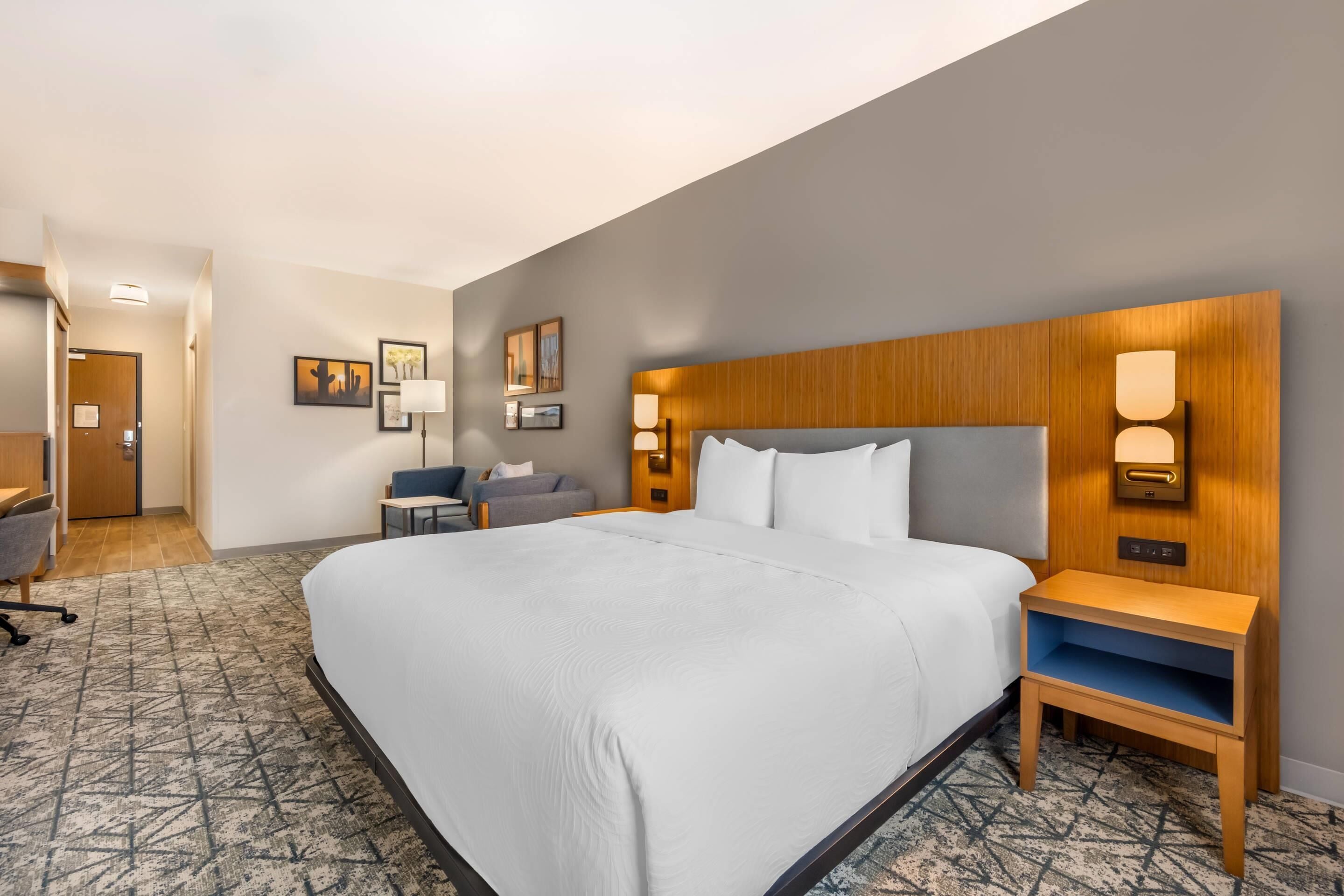 A hotel room with a large bed, a desk, a chair, a nightstand, and a wall-mounted lamp.