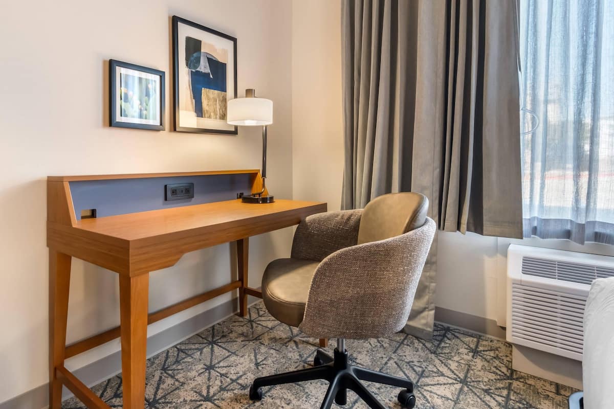 A hotel room with a wooden desk, a cushioned chair, a lamp, framed pictures on the wall, and an air conditioning unit.