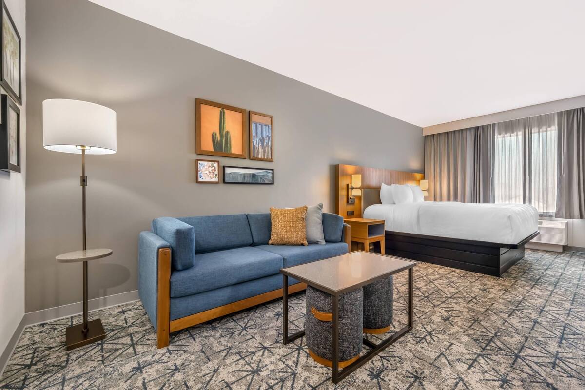 A hotel room with a blue sofa, a bed, a lamp, and framed artwork on the wall.