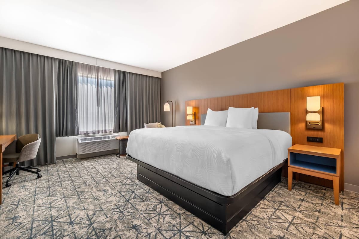 A hotel room with a large bed, a desk, a chair, a heater, and a window with curtains.