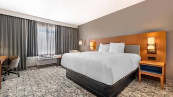 A hotel room with a large bed, a desk, a chair, a heater, and a window with curtains.